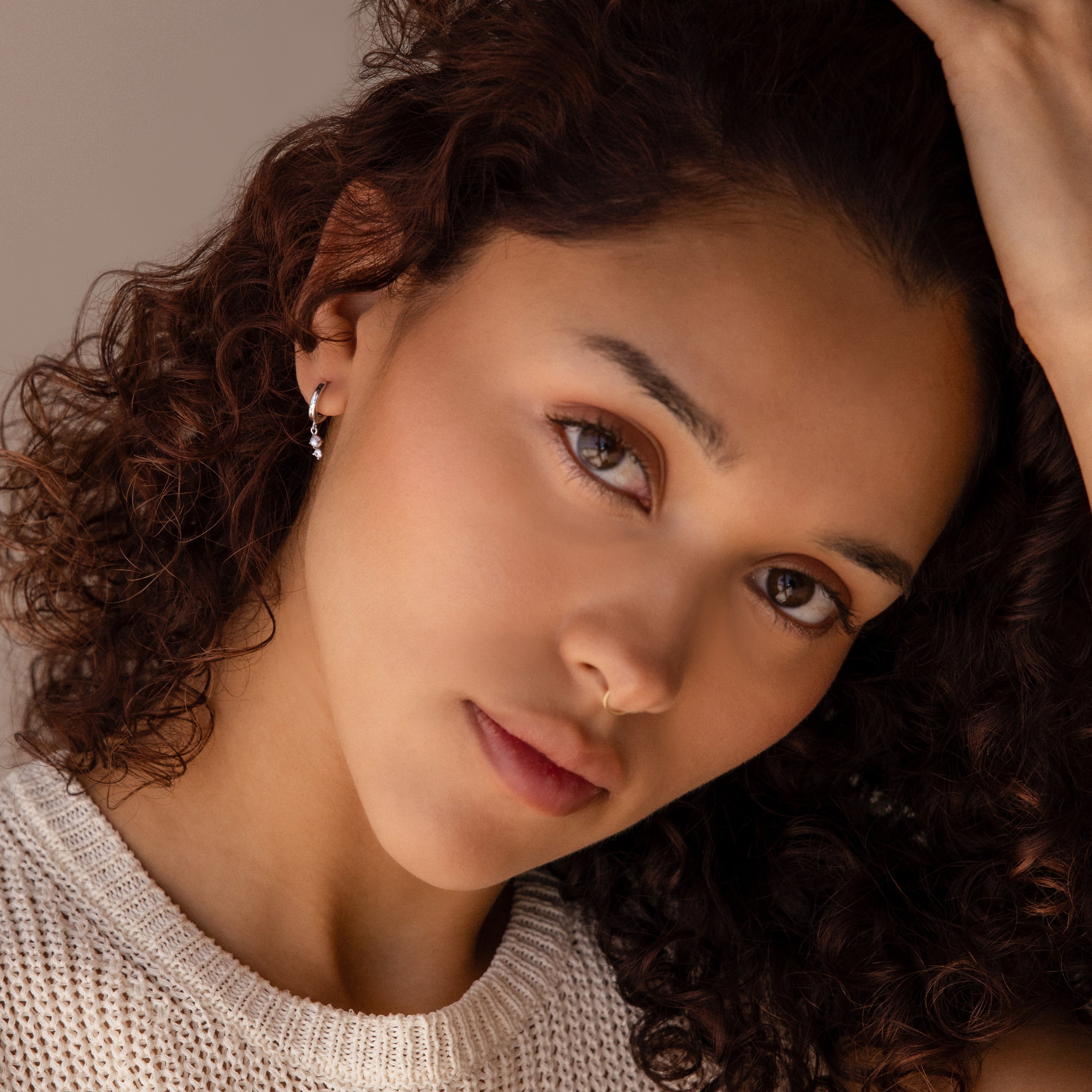 Woman with curly hair and a neutral expression wears a light knit top, looking to the side. Her look is complemented by Dangling Pearl Diamond Huggies in Sterling Silver—an elegant jewelry choice.