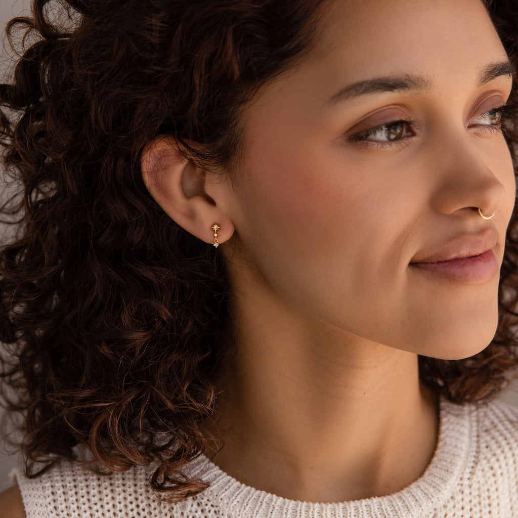 Woman with curly brown hair wears Diamond Starburst Drop Studs in 18K Gold and a nose ring, paired with a white knit top and a soft smile.