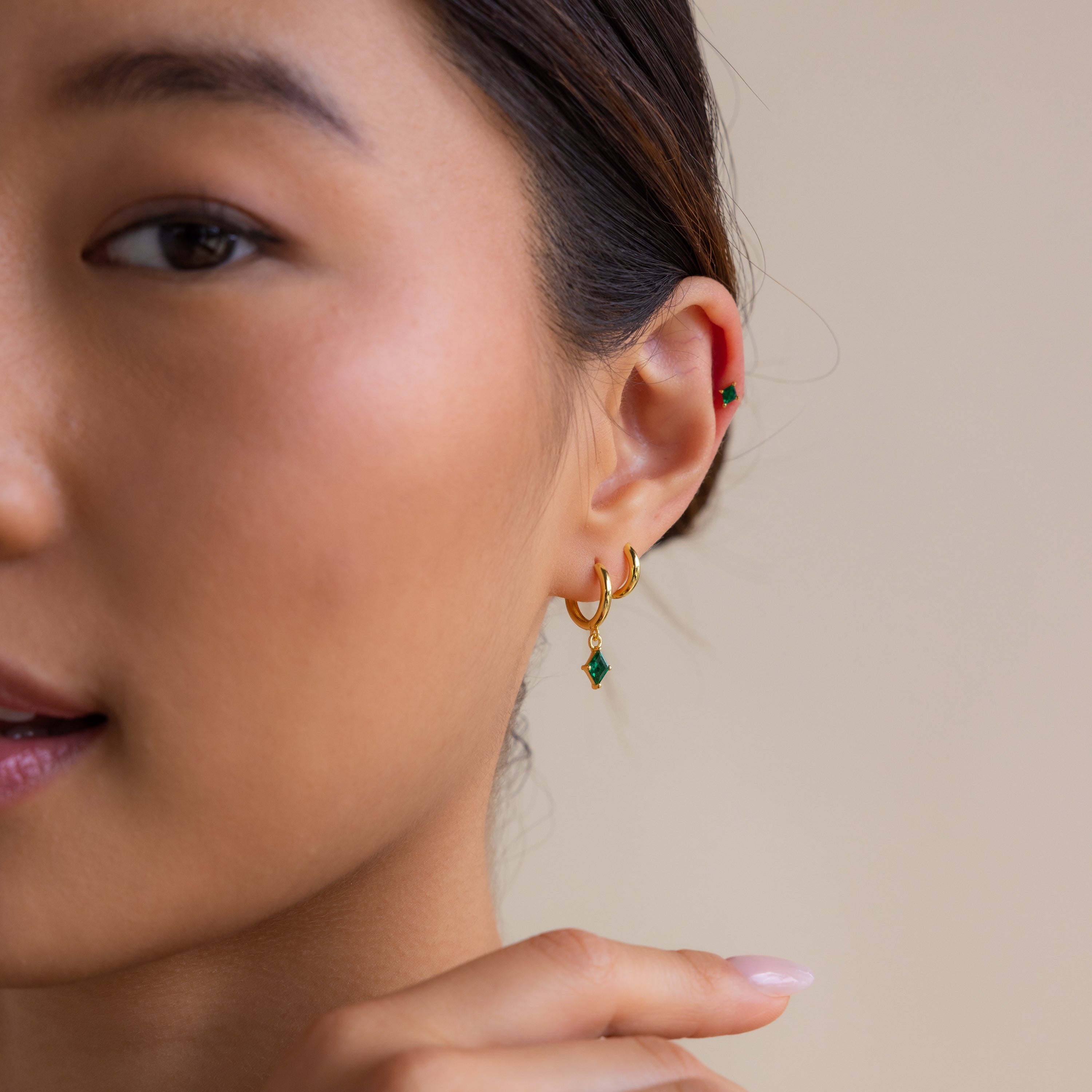 A woman wearing the Emerald Earrings Set, with gold hoops and green gems, touches her ear against a neutral beige background.