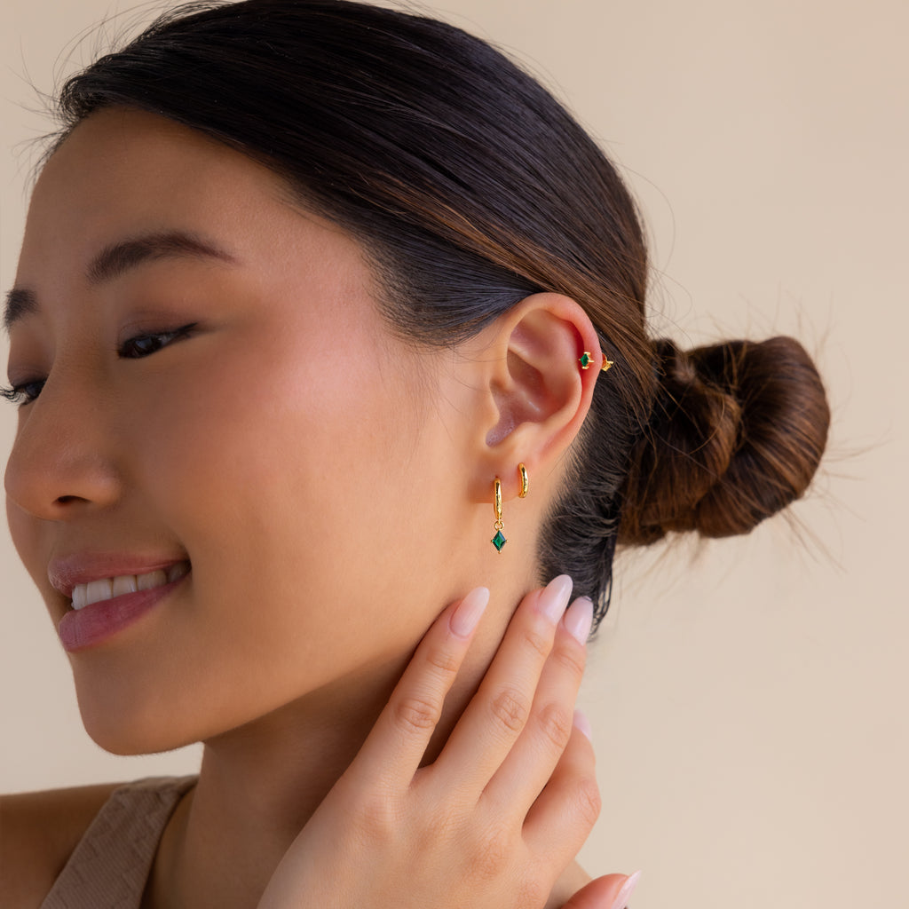 A woman with a neat bun, smiling as she gently touches her ear, showcases the elegant Emerald Earrings Set in gold and green.