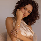 Woman with curly hair poses in a satin top, wearing gold jewelry with pearls and the Hinged Circle Charm Bracelet in 18K Gold, looking at the camera.