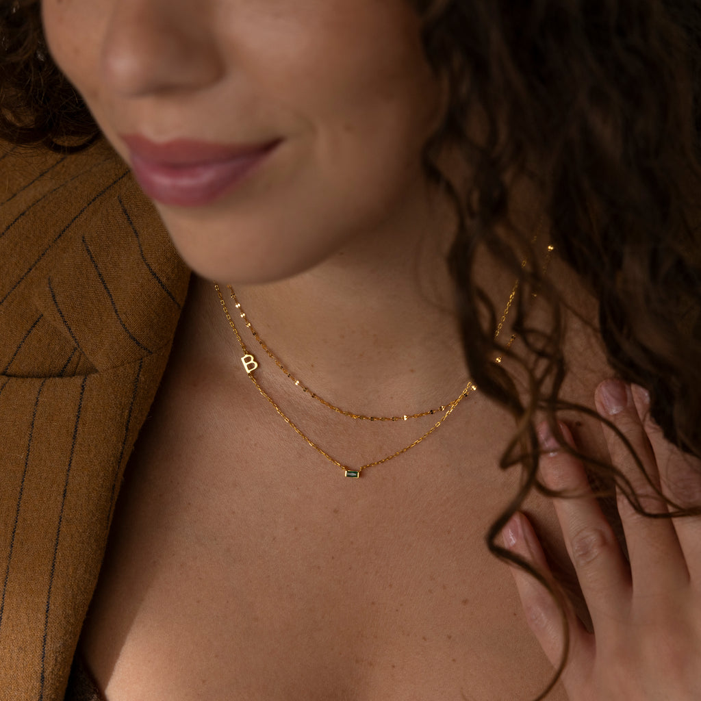 Smiling woman in a brown pinstripe blazer wears a gold layered set featuring the Initial Baguette Birthstone Necklace with a "B" letter, paired with a sparkling sideways initial necklace.