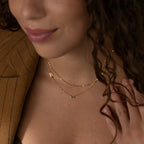 Smiling woman in a brown pinstripe blazer wears a gold layered set featuring the Initial Baguette Birthstone Necklace with a "B" letter, paired with a sparkling sideways initial necklace.