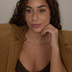 A woman with curly hair in a brown pinstripe blazer wears an Initial Baguette Birthstone Necklace and smiles gently, resting her chin on her hand.