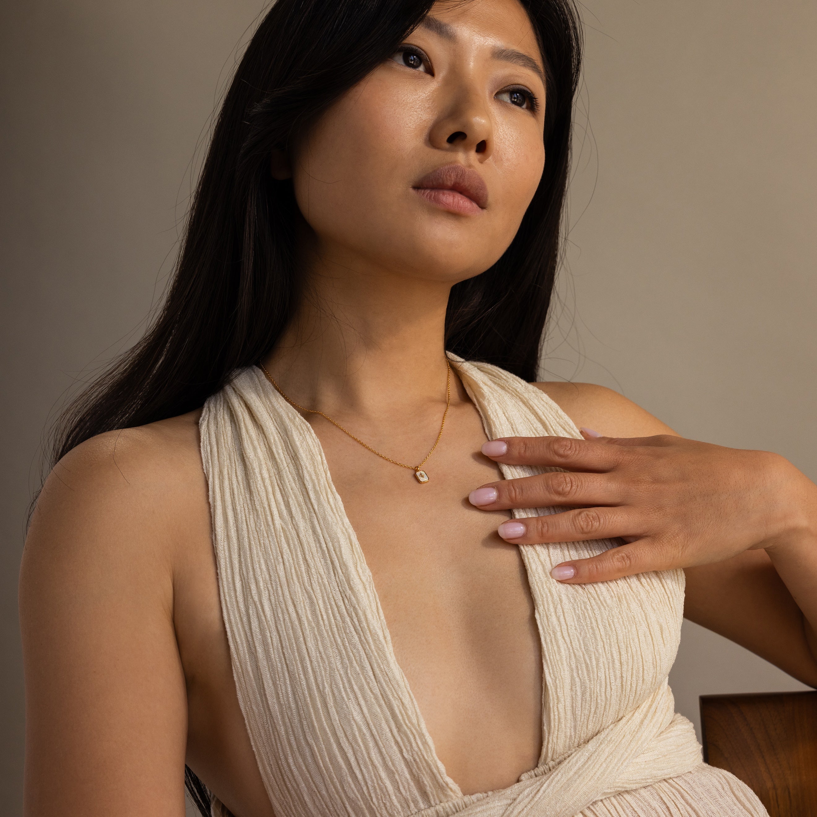 A woman with long dark hair in a cream textured dress touches her North Star Opal Necklace, gazing slightly upward.