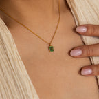 The North Star Emerald Necklace, featuring a green crystal pendant, is worn around the neck of someone with light pink polished nails.