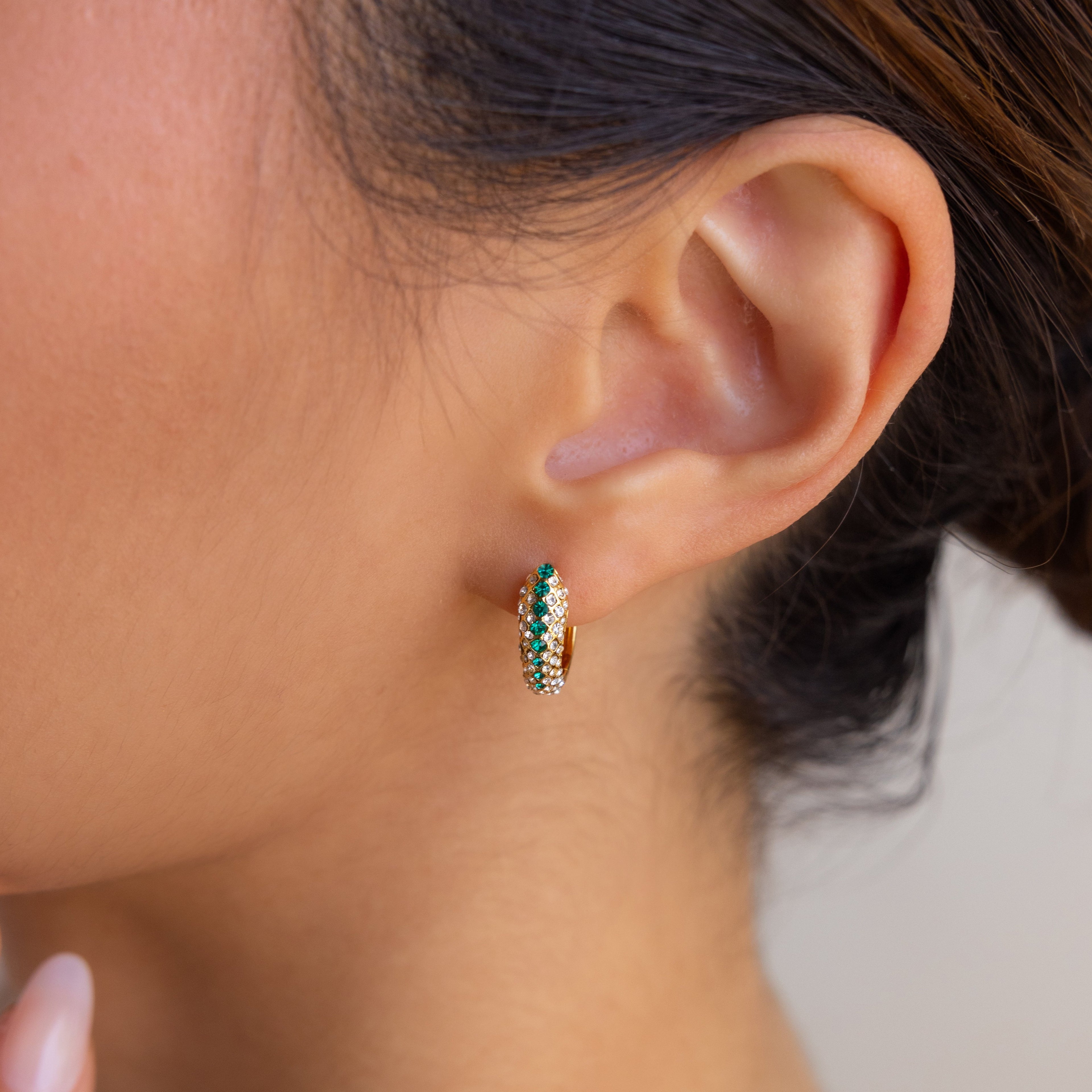 A close-up shows a woman's ear adorned with Emerald Pave Snake Hoops—gold hoops accented by green and white stones, featuring a subtle snakeskin texture.