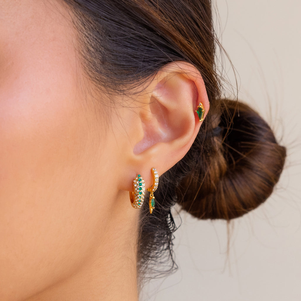 A close-up of a woman's ear wearing Emerald Pave Snake Hoops, highlighting their elegant snakeskin texture and sparkling emerald details.