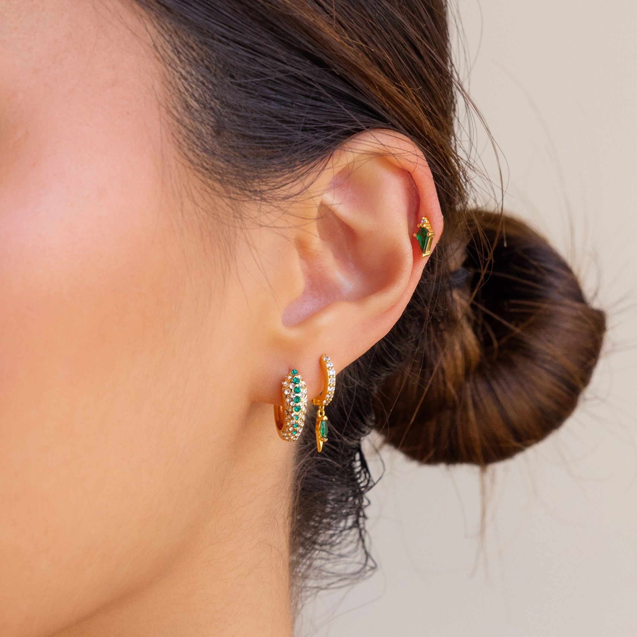 A close-up of a woman's ear wearing Emerald Pave Snake Hoops, highlighting their elegant snakeskin texture and sparkling emerald details.