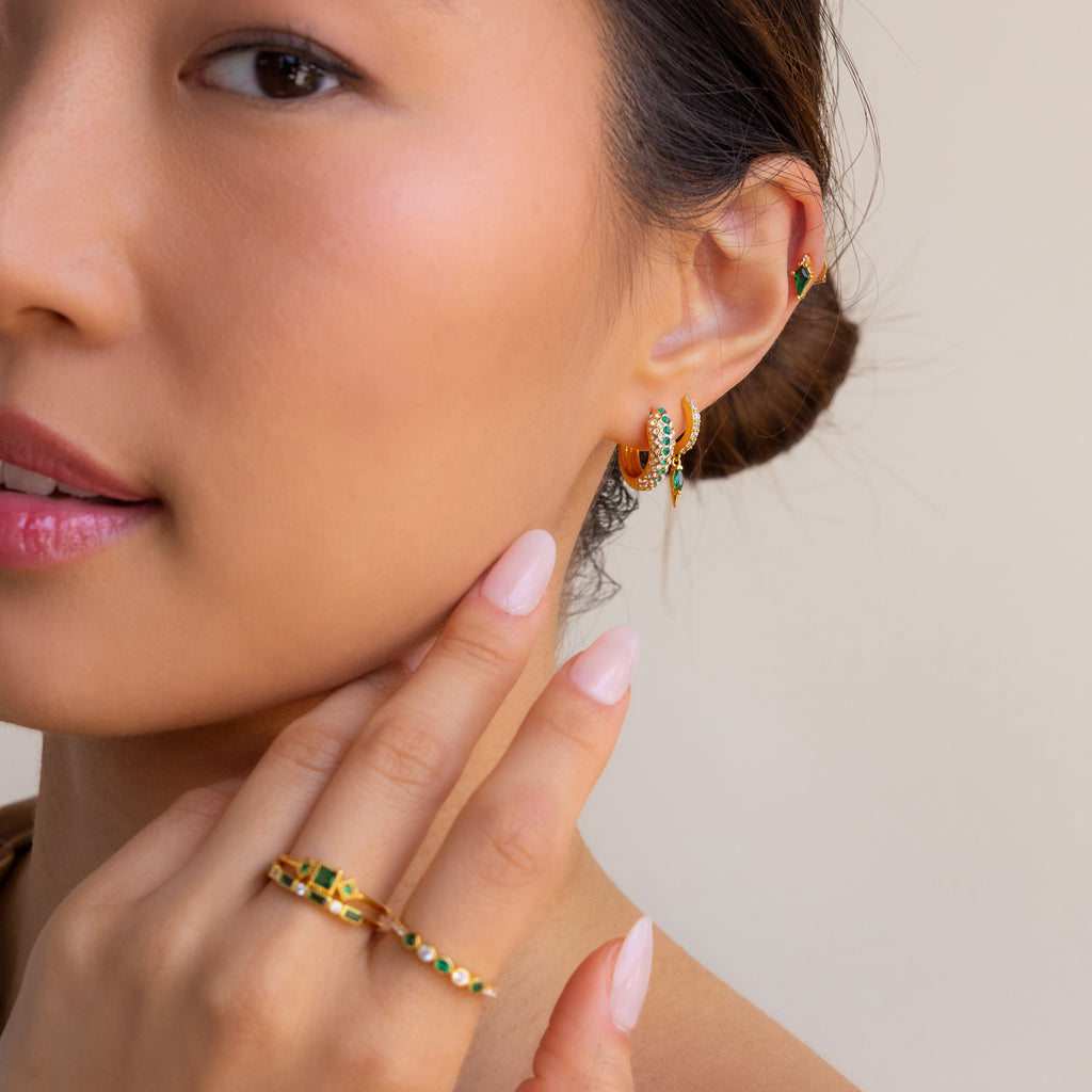 A woman with manicured nails gently touches her face while wearing the Emerald Pave Snake Hoops, along with rings and an ear cuff.