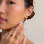 A woman with manicured nails gently touches her face while wearing the Emerald Pave Snake Hoops, along with rings and an ear cuff.