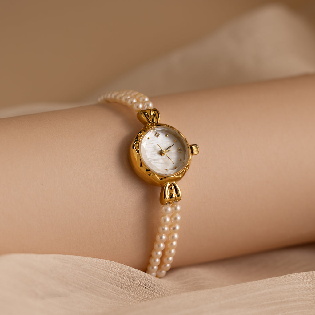 The Beaded Pearl Timepiece in 18K Gold, featuring a gold face and elegant pearl strap, adorns a wrist against a soft beige fabric backdrop.