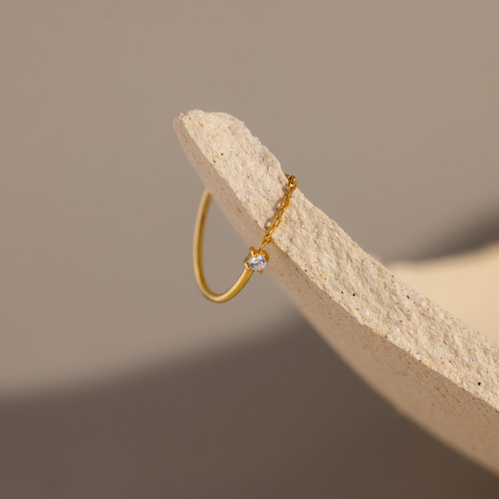 The Diamond Half Chain Ring, a delicate gold piece with sparkling diamonds, is styled with dainty jewelry and set on the edge of a textured beige surface.