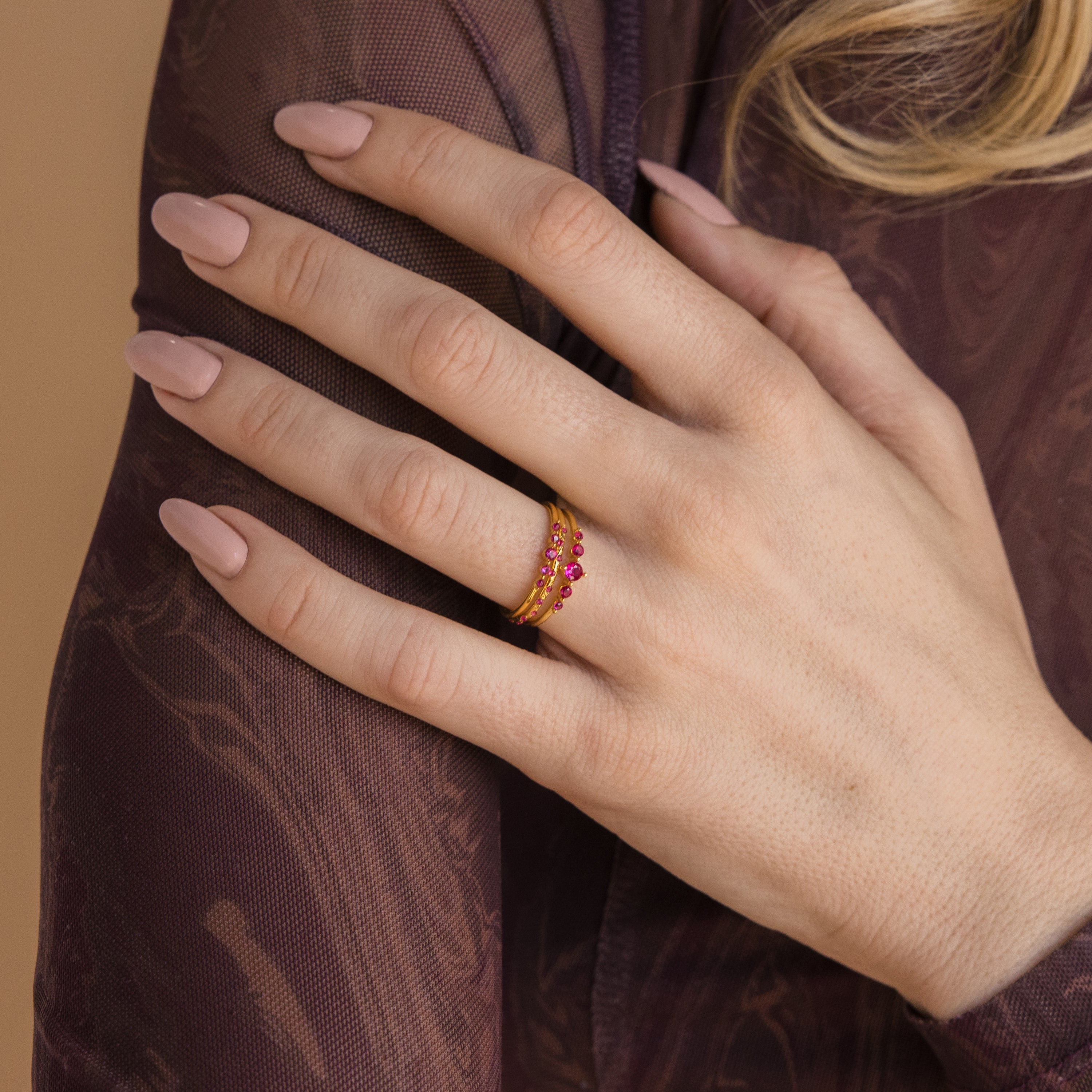 A hand with nude nail polish wears the Lace Birthstone Rings Set, featuring gold minimalist rings with red stones, resting on a purple sheer-sleeved arm.