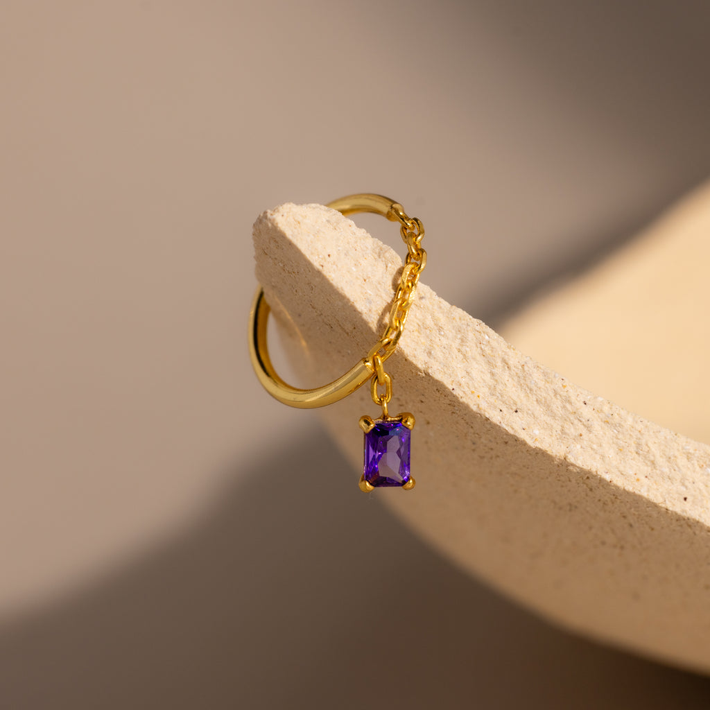 The Amethyst Drop Chain Ring, featuring a delicate gold chain and a small purple gemstone charm, rests on a textured beige surface.