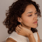 Woman with curly hair wearing a sleeveless knit top, touching her Pearl Ivy Flower Huggies in Sterling Silver, and looking to the side.