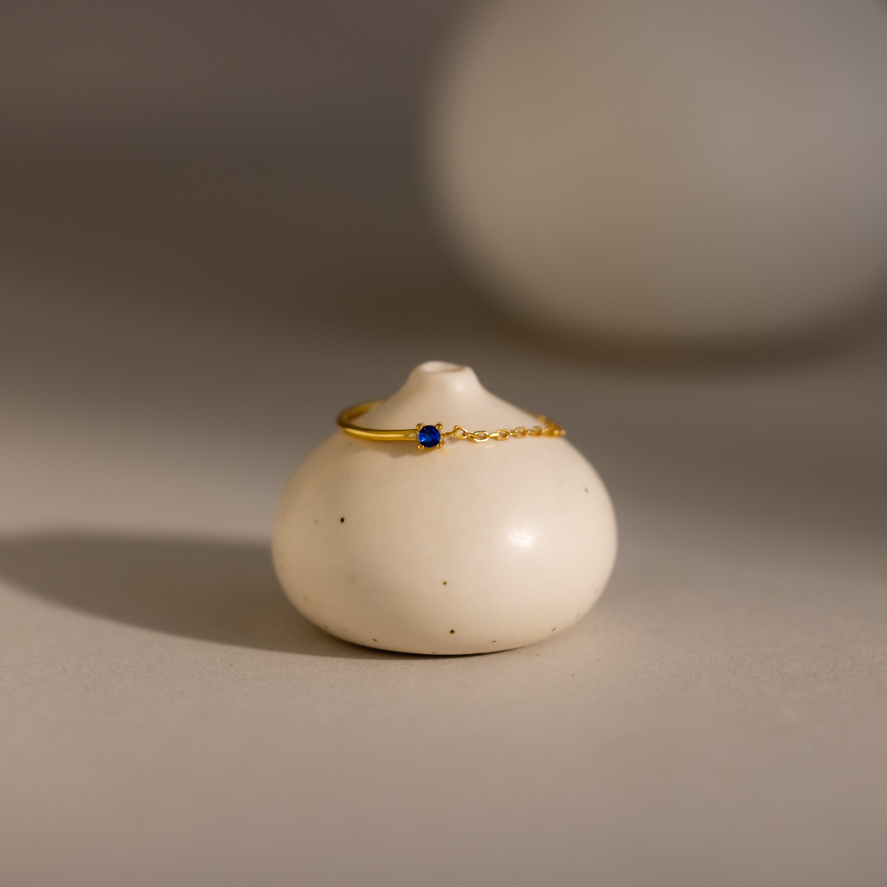 The Sapphire Half Chain Ring, featuring a delicate gold band with a small blue gemstone, is perfect for dainty jewelry lovers and is elegantly displayed on a round, white ceramic vase.