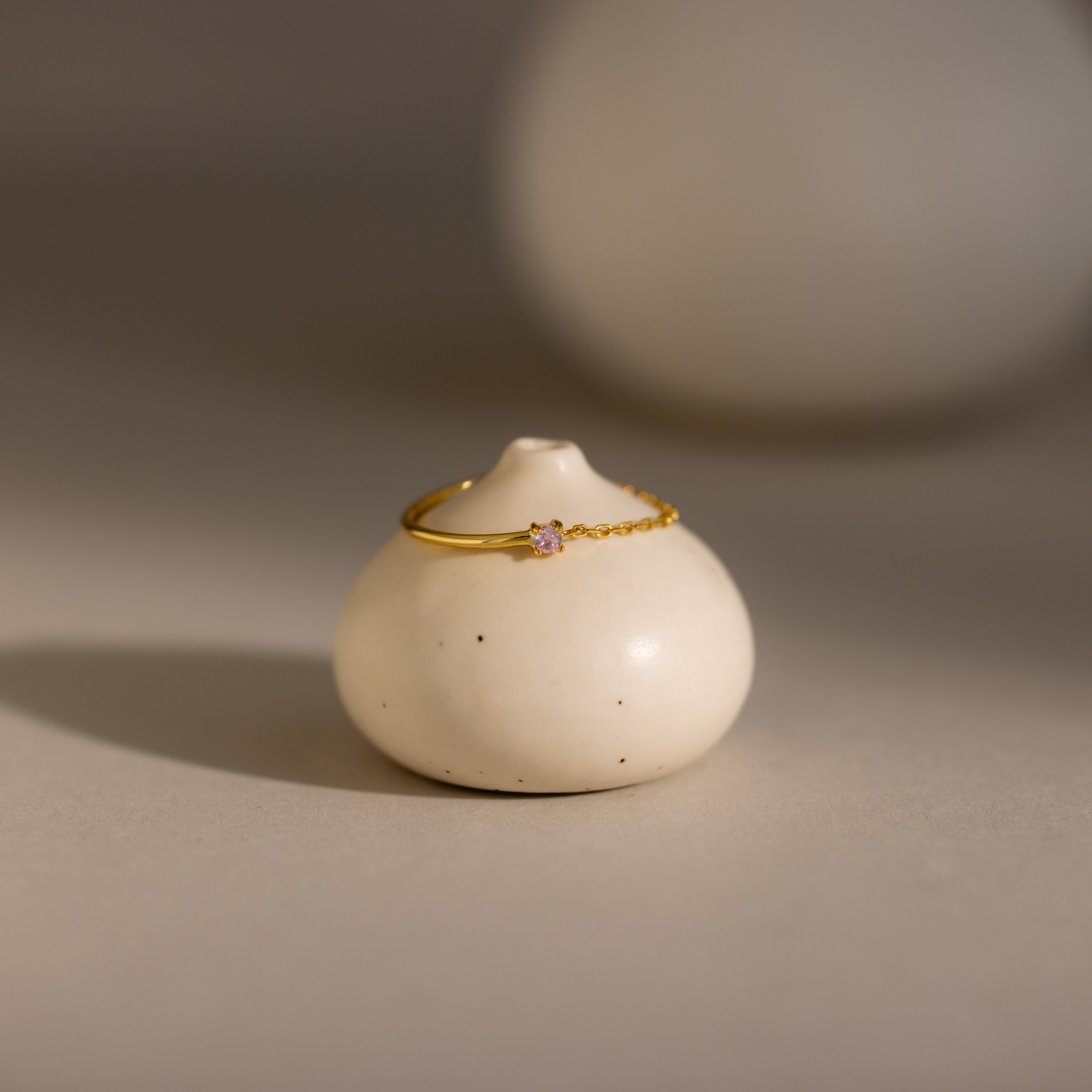 The Pink Tourmaline Half Chain Ring features a delicate gold band and a small pink stone, elegantly displayed on a round white ceramic vase—a perfect piece of dainty jewelry.