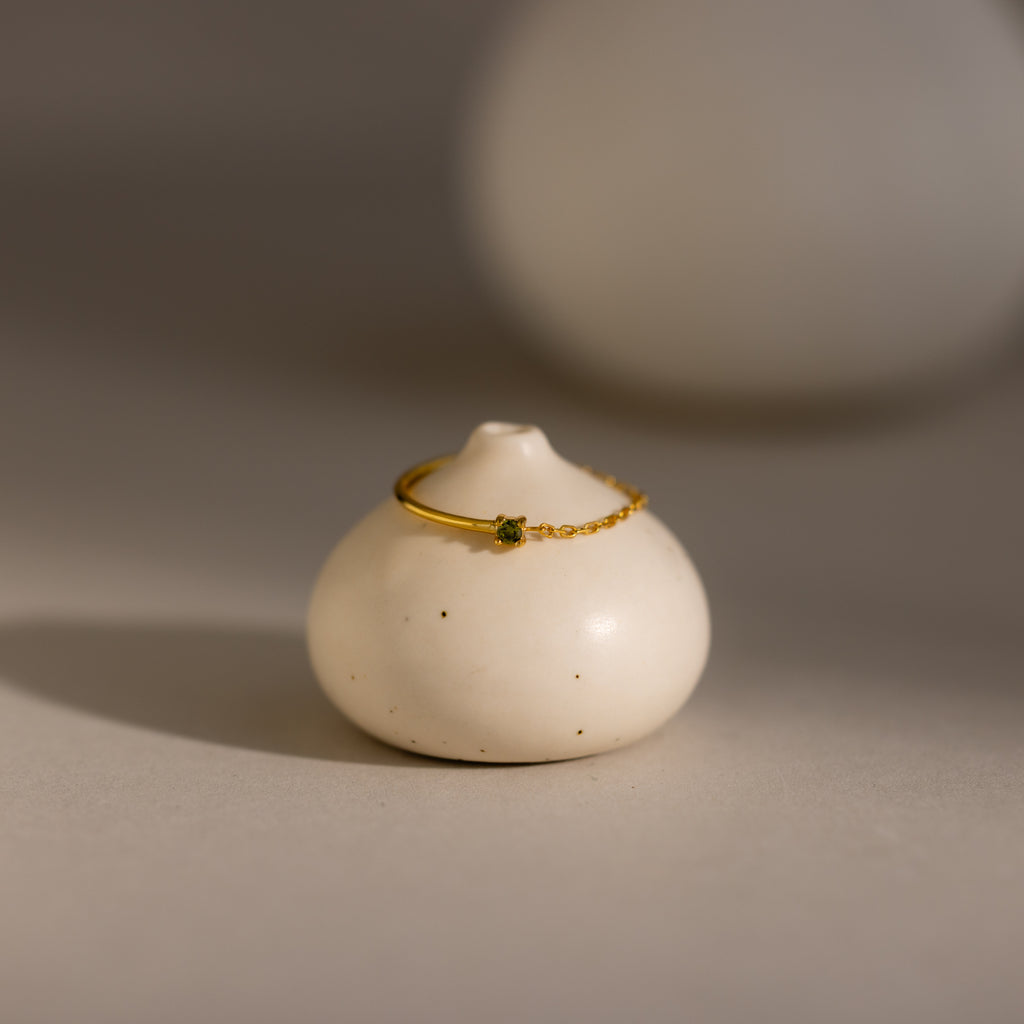 The Peridot Half Chain Ring is a delicate gold ring with a small peridot stone, shown on a smooth, round white ceramic holder—ideal for those who love dainty jewelry.