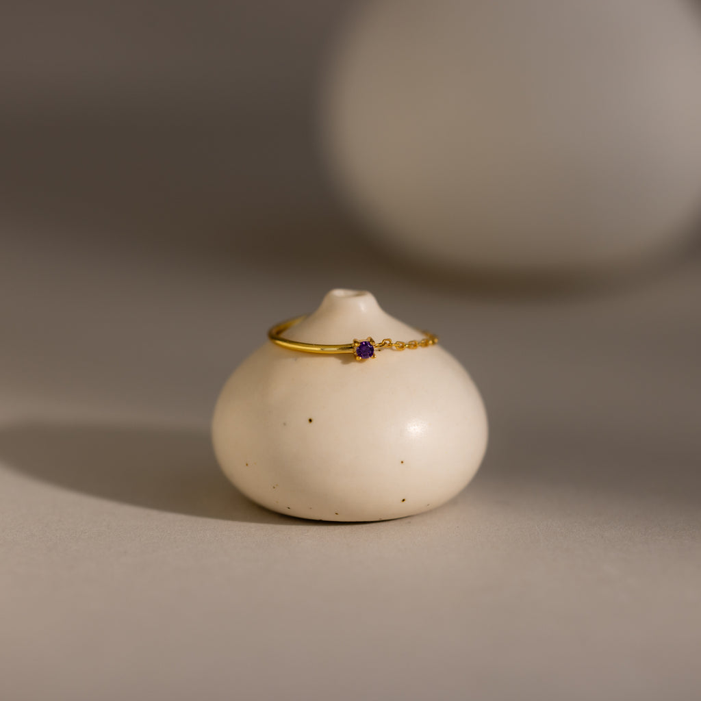 The Amethyst Half Chain Ring, featuring a delicate purple stone set in gold, is displayed on a round white ceramic holder against a soft background.