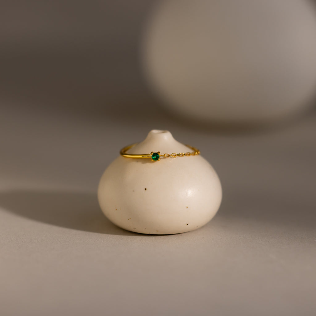 The Emerald Half Chain Ring, a delicate gold piece with a small green gemstone, is perfect as a birthstone ring and is displayed on a round white ceramic object.