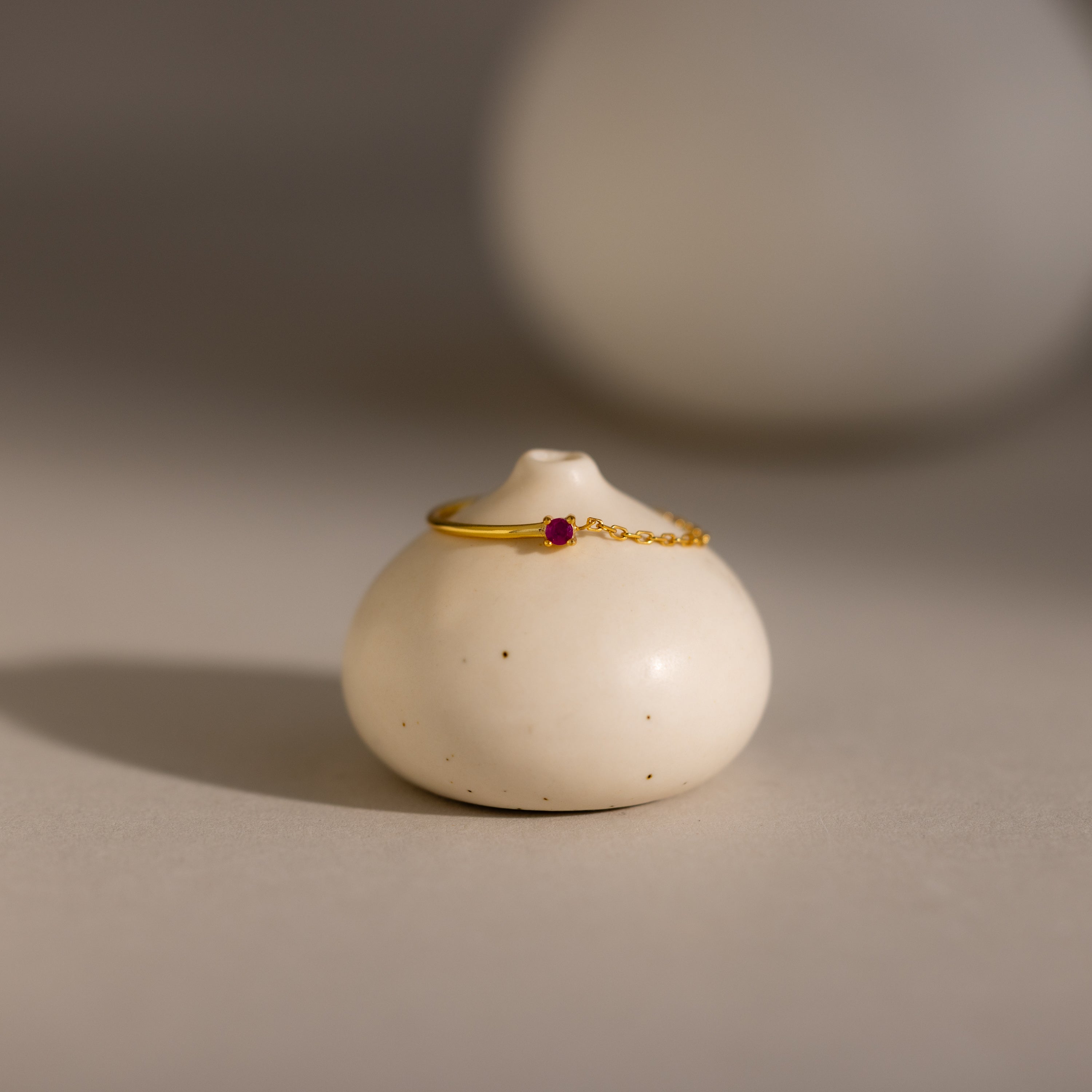 The Ruby Half Chain Ring is a dainty gold jewelry piece with a small red gemstone, beautifully displayed on a round white ceramic stand under soft lighting.