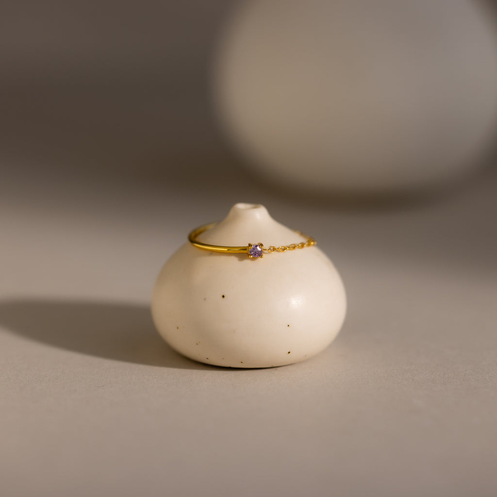 The Alexandrite Half Chain Ring, a delicate gold band with a petite gem, is displayed on a smooth round white ceramic holder—ideal for fans of dainty jewelry.