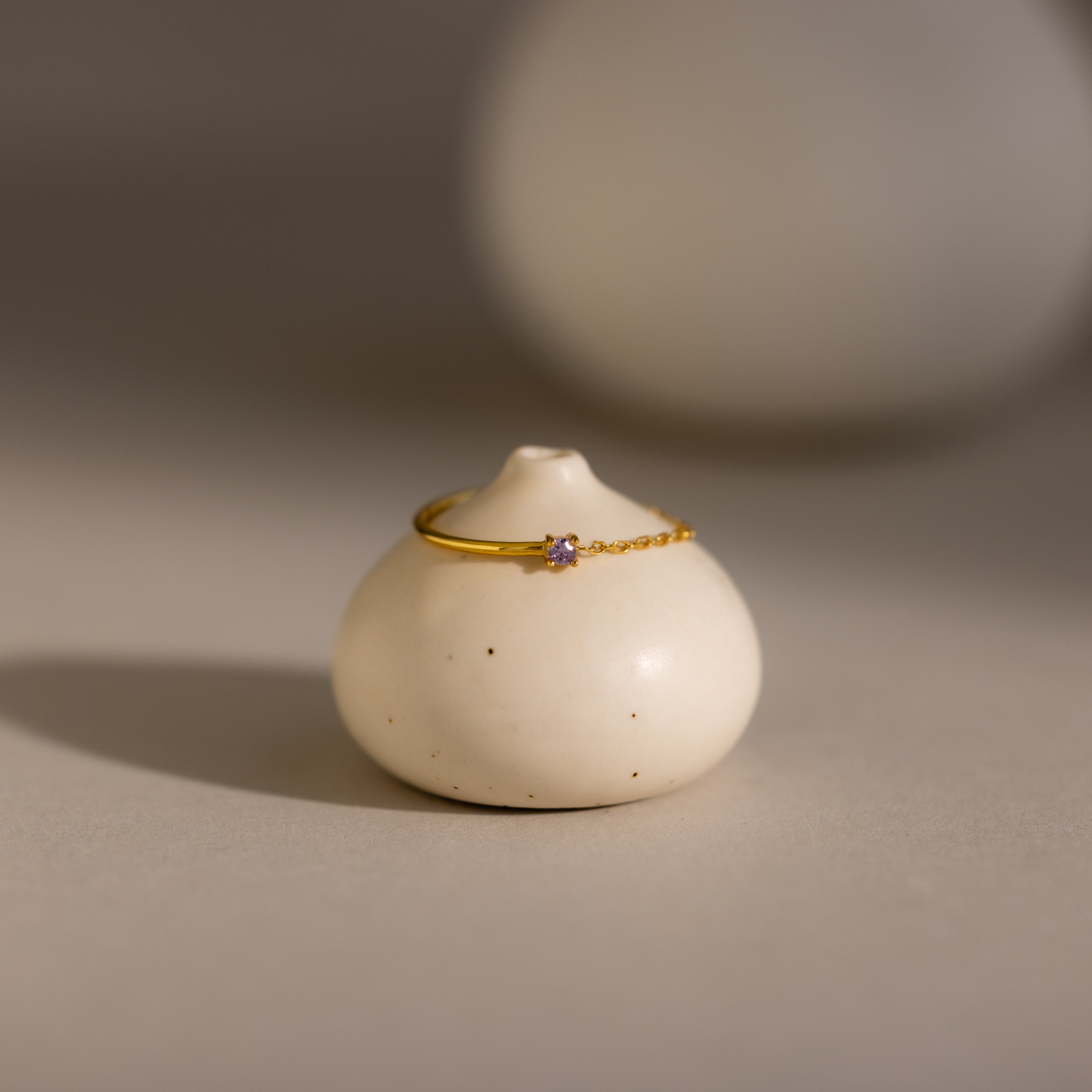 The Alexandrite Half Chain Ring, a delicate gold band with a petite gem, is displayed on a smooth round white ceramic holder—ideal for fans of dainty jewelry.