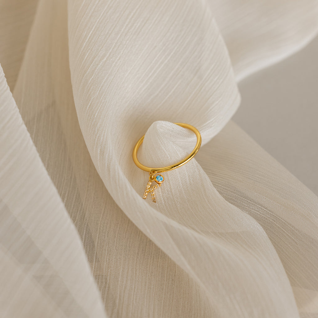 The Pave Initial Birthstone Drop Ring, featuring a blue gem and letter A charm, rests on soft cream fabric—a perfect customizable ring for a personal touch.