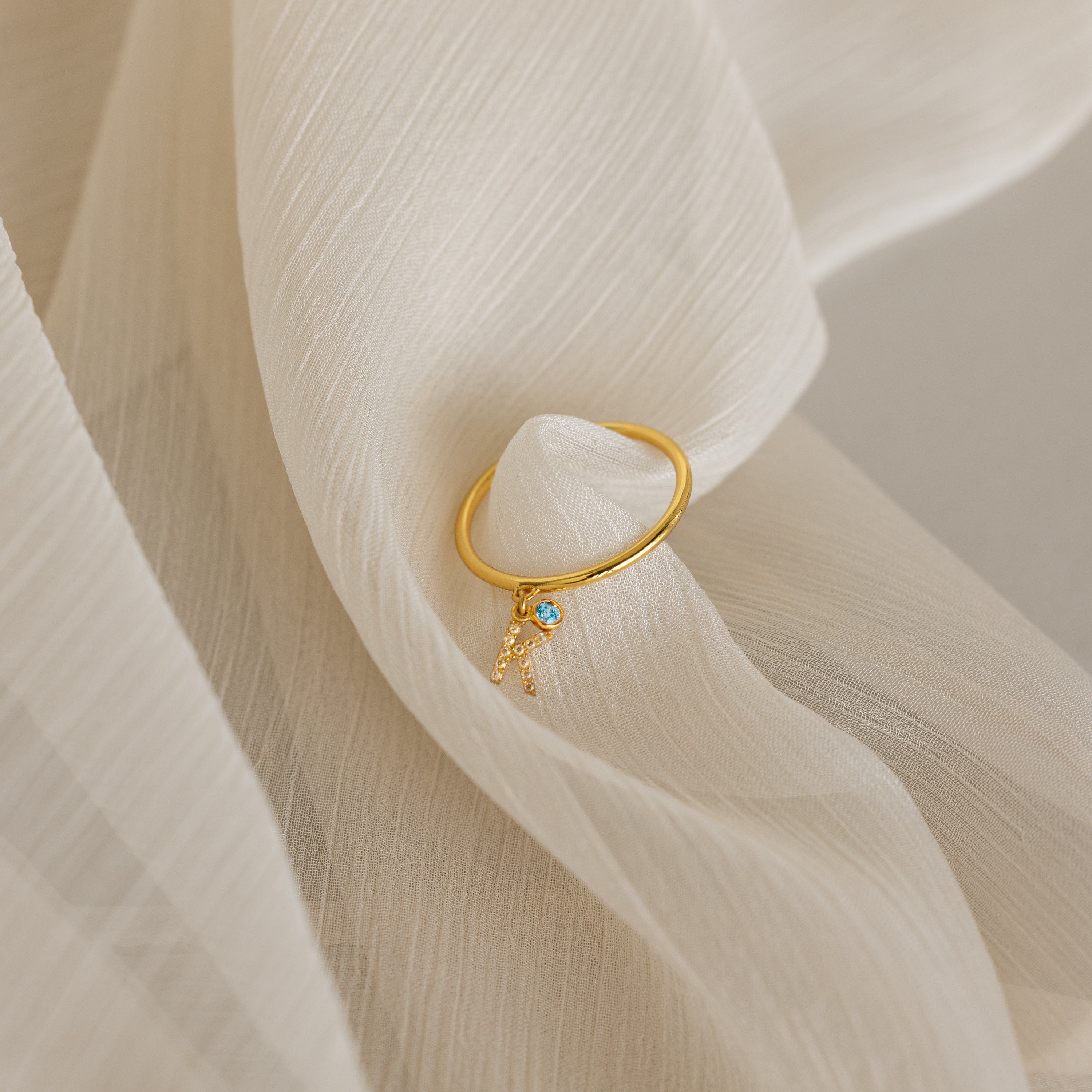 The Pave Initial Birthstone Drop Ring, featuring a blue gem and letter A charm, rests on soft cream fabric—a perfect customizable ring for a personal touch.