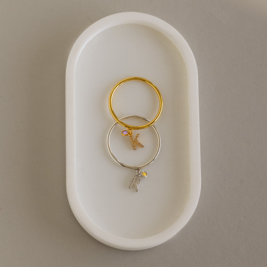 Two Pave Initial Birthstone Drop Rings—one gold, one silver—each with a dangling letter K charm, rest on a white oval dish. Perfect as charm rings or for customizing with your chosen initial and birthstone.
