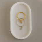 Two Pave Initial Birthstone Drop Rings—one gold, one silver—each with a dangling letter K charm, rest on a white oval dish. Perfect as charm rings or for customizing with your chosen initial and birthstone.