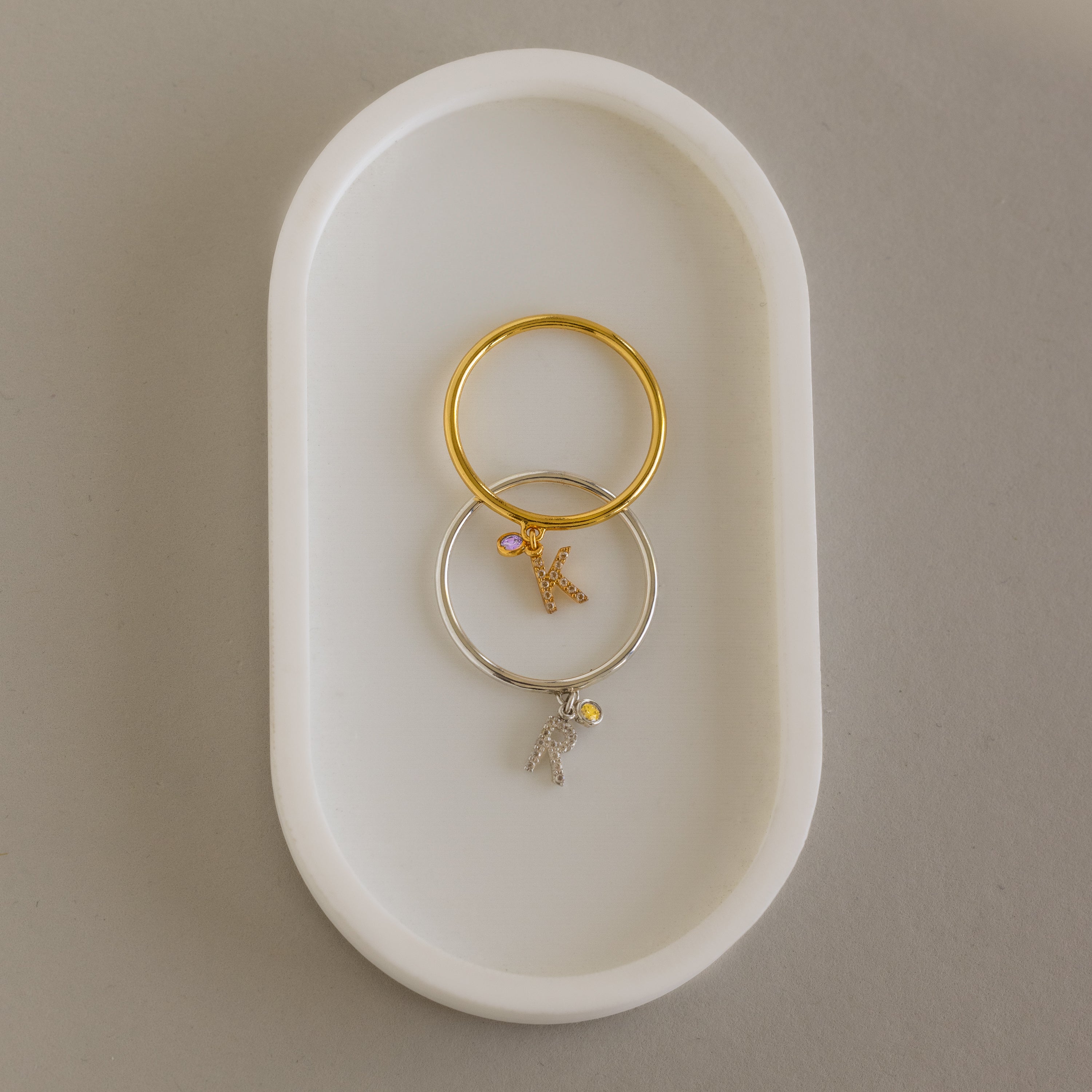 Two Pave Initial Birthstone Drop Rings—one gold, one silver—each with a dangling letter K charm, rest on a white oval dish. Perfect as charm rings or for customizing with your chosen initial and birthstone.