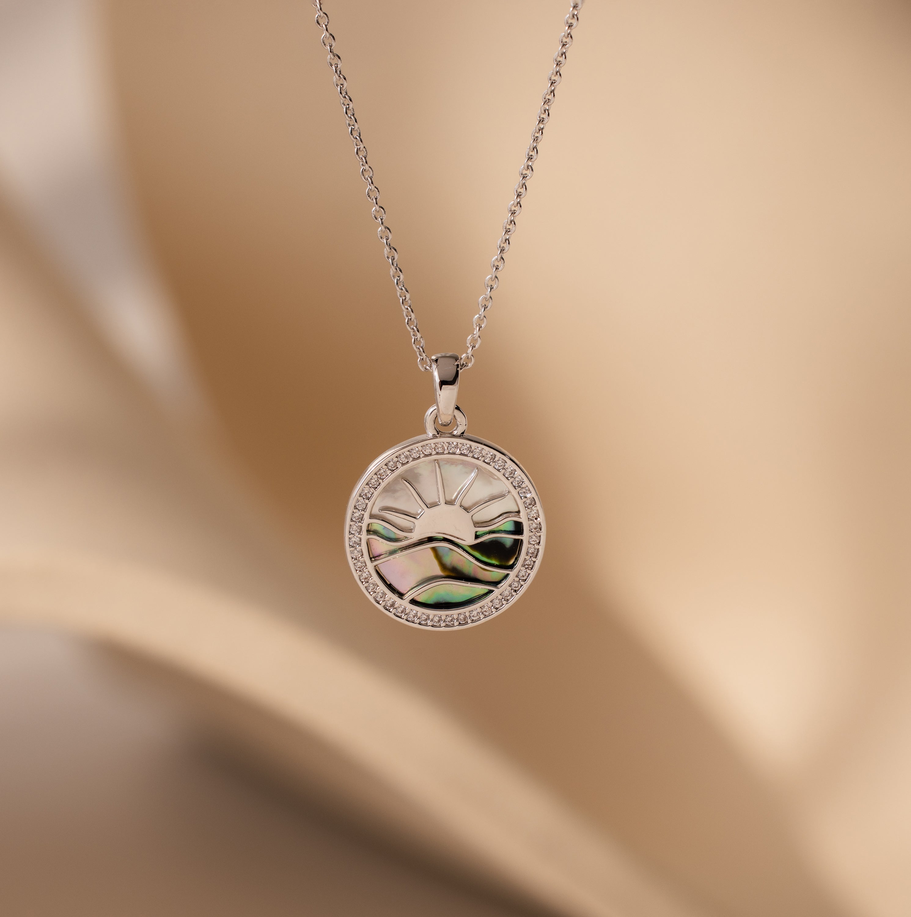 Abalone Sunrise Pendant Necklace in White Gold featuring a sun and landscape design with shimmering abalone and pearl gemstones on a soft beige background.