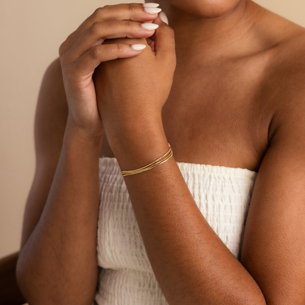 A woman in a white textured top wears the Triple Snake Chain Bracelet in 18K Gold on her wrist, embracing minimal jewelry for an effortlessly chic look with gently clasped hands.