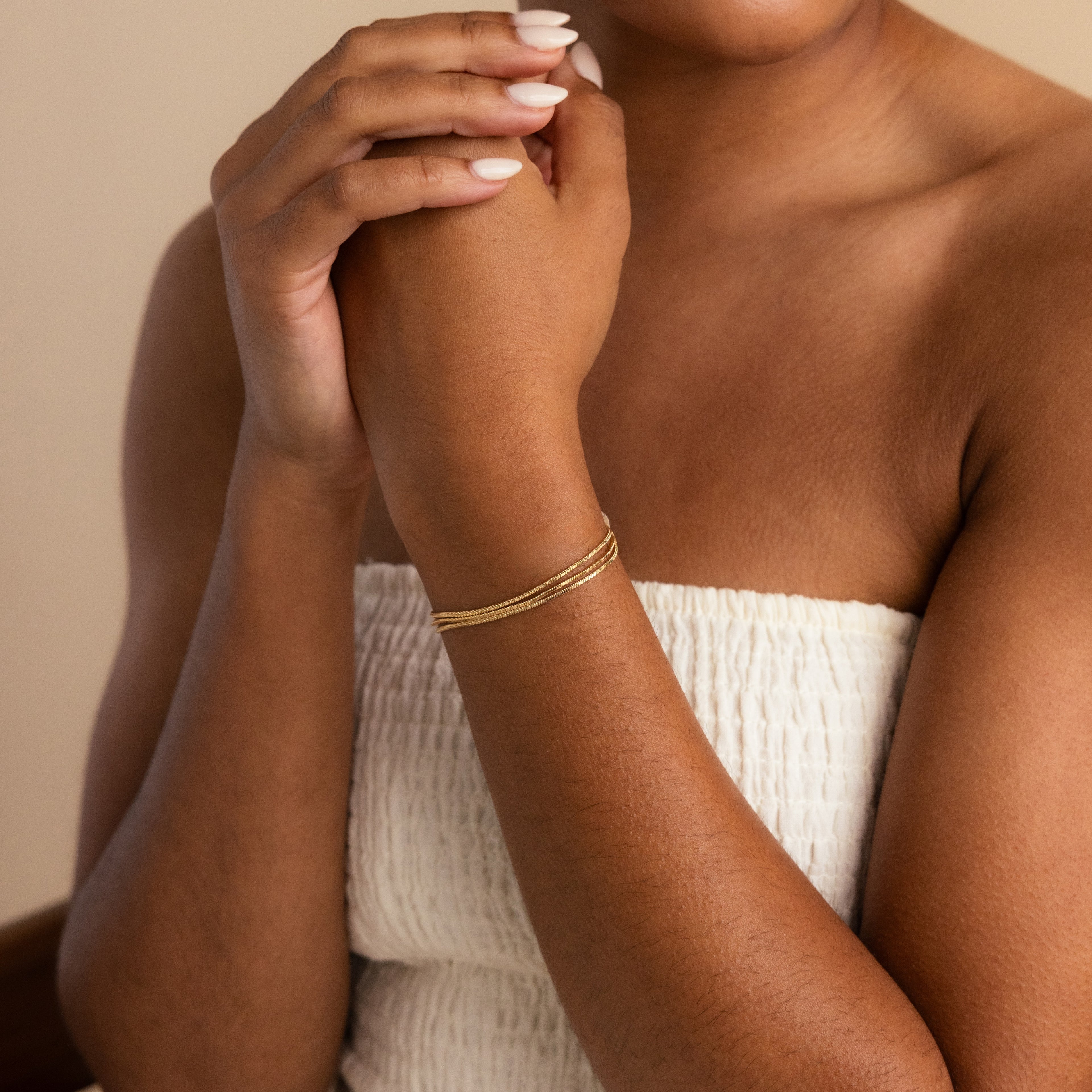 A woman in a white textured top wears the Triple Snake Chain Bracelet in 18K Gold on her wrist, embracing minimal jewelry for an effortlessly chic look with gently clasped hands.