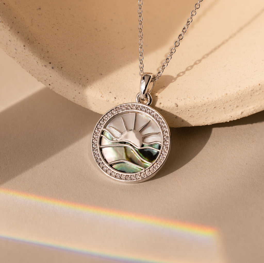 The Abalone Sunrise Pendant Necklace in White Gold, featuring a sun and landscape design with abalone and pearl gemstones, is displayed on a neutral background with a rainbow light.