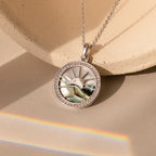 The Abalone Sunrise Pendant Necklace in White Gold, featuring a sun and landscape design with abalone and pearl gemstones, is displayed on a neutral background with a rainbow light.