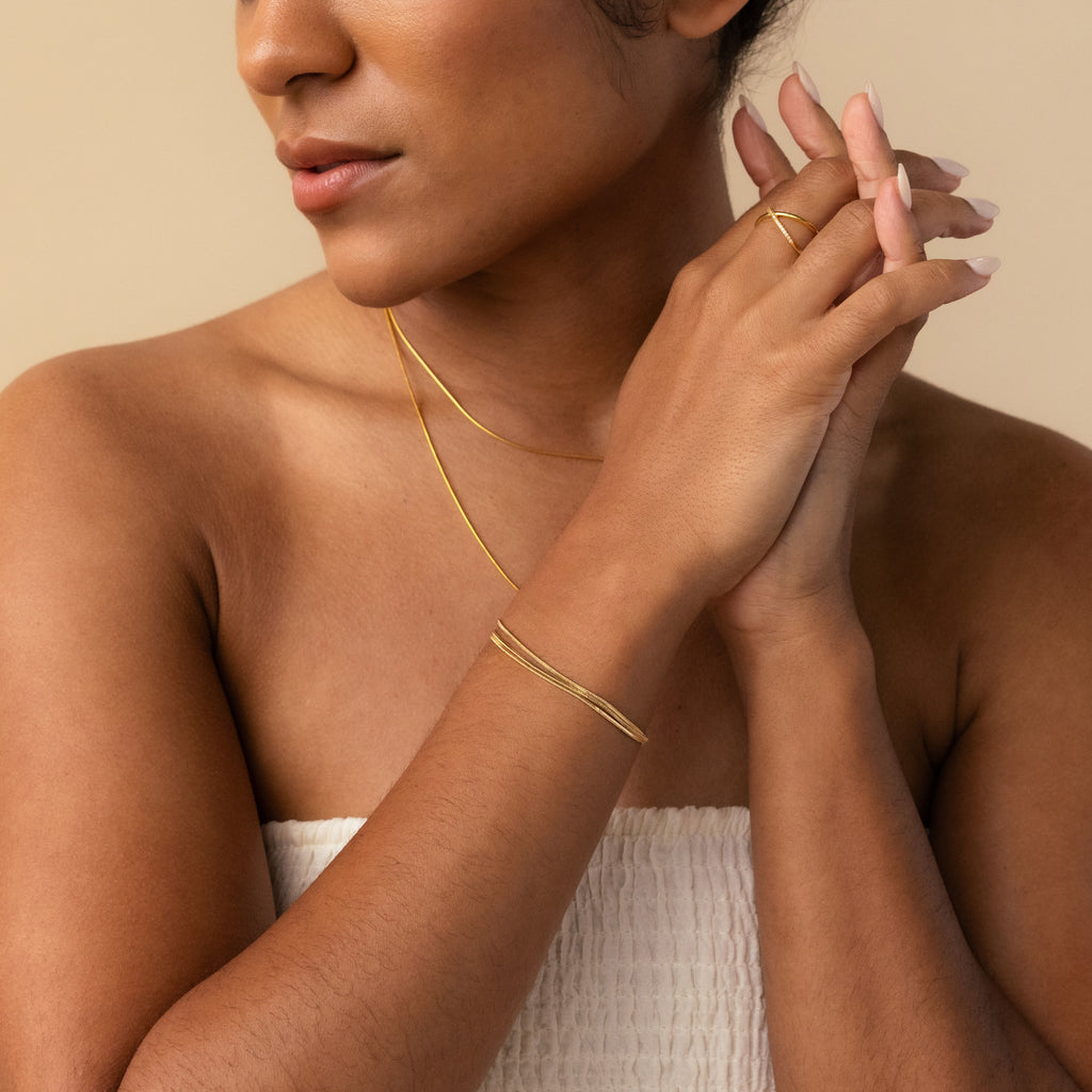 A woman with nude nails and minimal jewelry—a gold necklace, the Triple Snake Chain Bracelet in 18K Gold, and a ring—stands against a beige background.