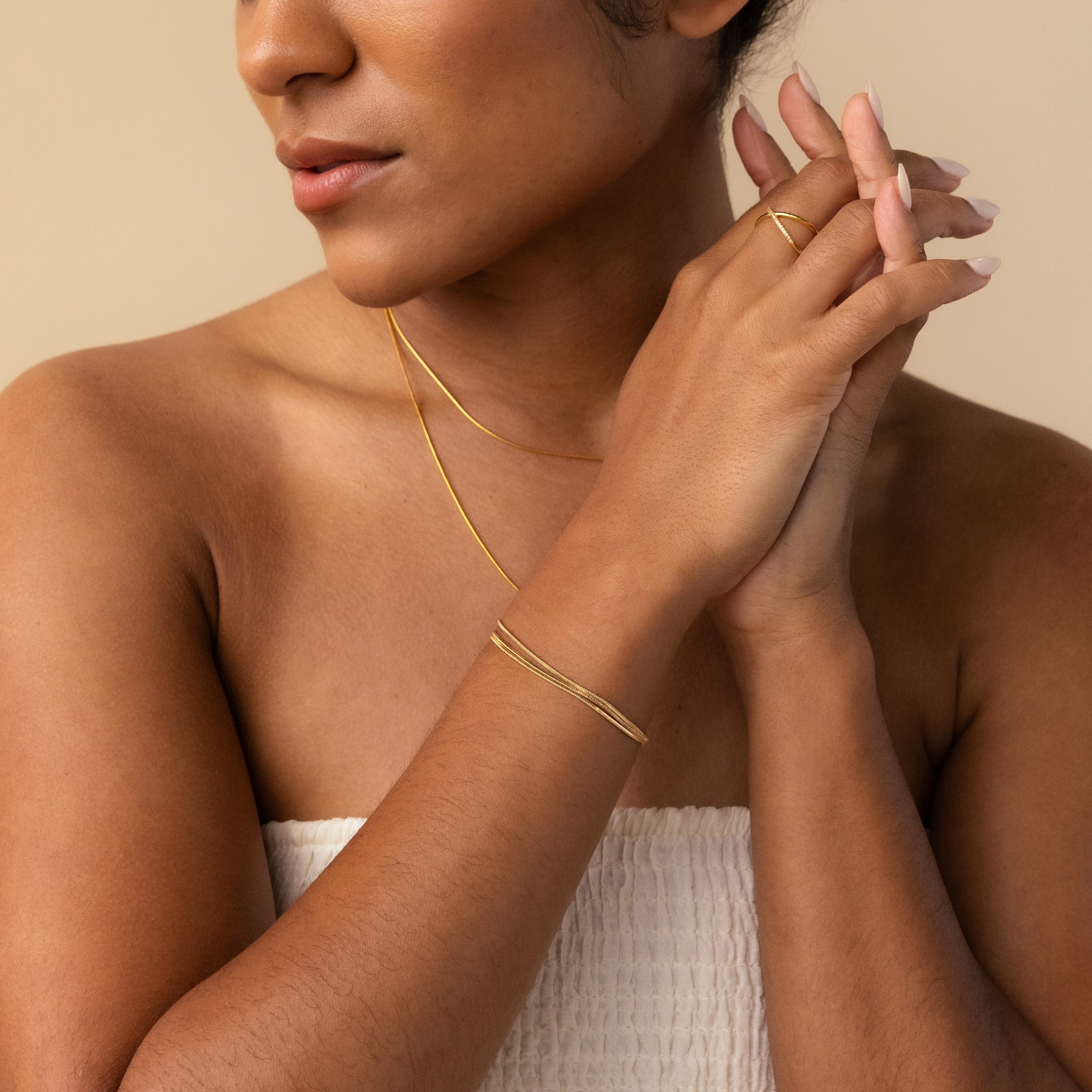 A woman with nude nails and minimal jewelry—a gold necklace, the Triple Snake Chain Bracelet in 18K Gold, and a ring—stands against a beige background.
