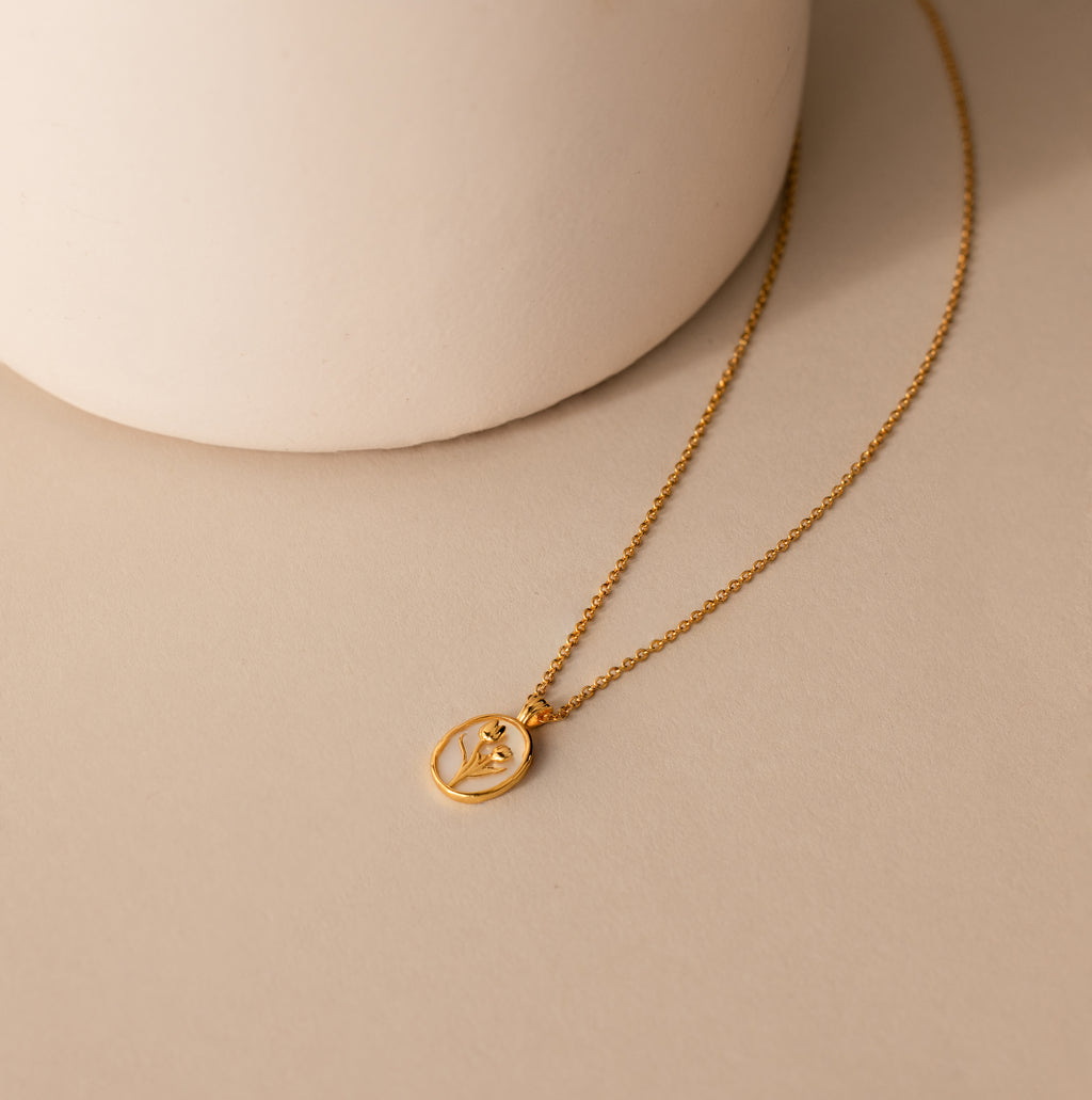 The Pearl Tulip Necklace in 18K Gold features a delicate leaf-inspired design, displayed on a beige surface beside a round white accent, blending the elegance of pearl jewelry with the charm of tulip motifs.