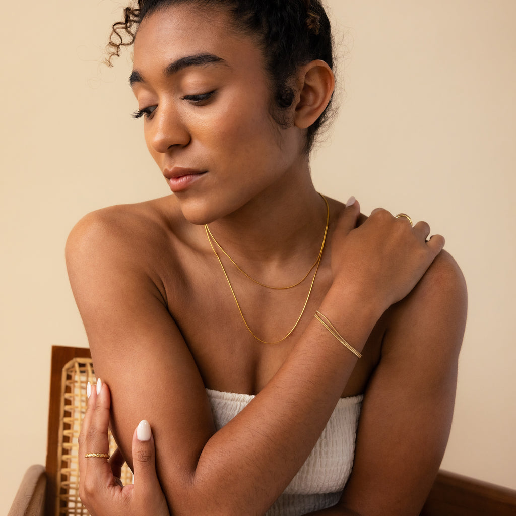 A woman in a white top styles her look with gold jewelry, featuring the Triple Snake Chain Bracelet for a layered effect, as she looks down with one arm crossed over her chest.
