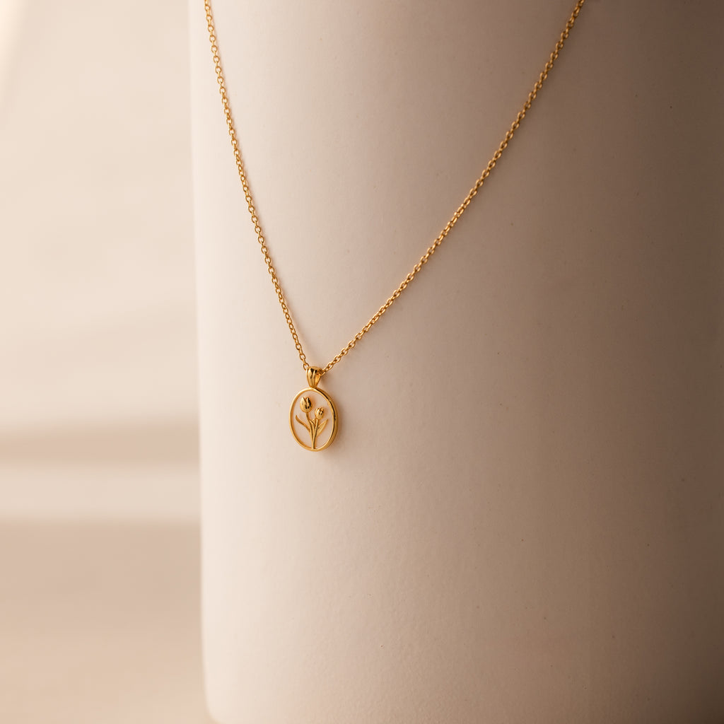 The Pearl Tulip Necklace in 18K Gold features an oval pendant with a minimalist plant design, elegantly displayed on a smooth beige surface.