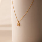 The Pearl Tulip Necklace in 18K Gold features an oval pendant with a minimalist plant design, elegantly displayed on a smooth beige surface.