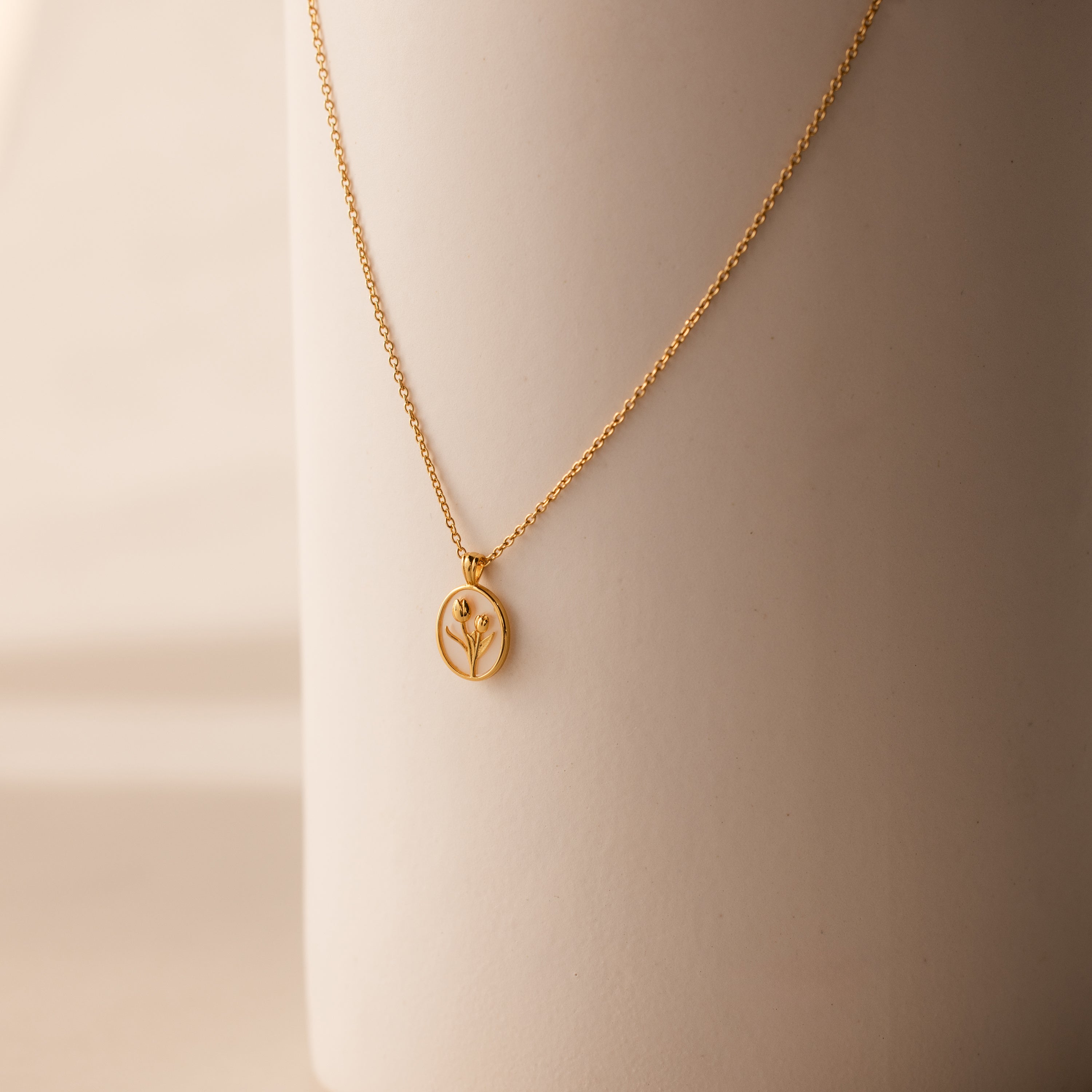 The Pearl Tulip Necklace in 18K Gold features an oval pendant with a minimalist plant design, elegantly displayed on a smooth beige surface.