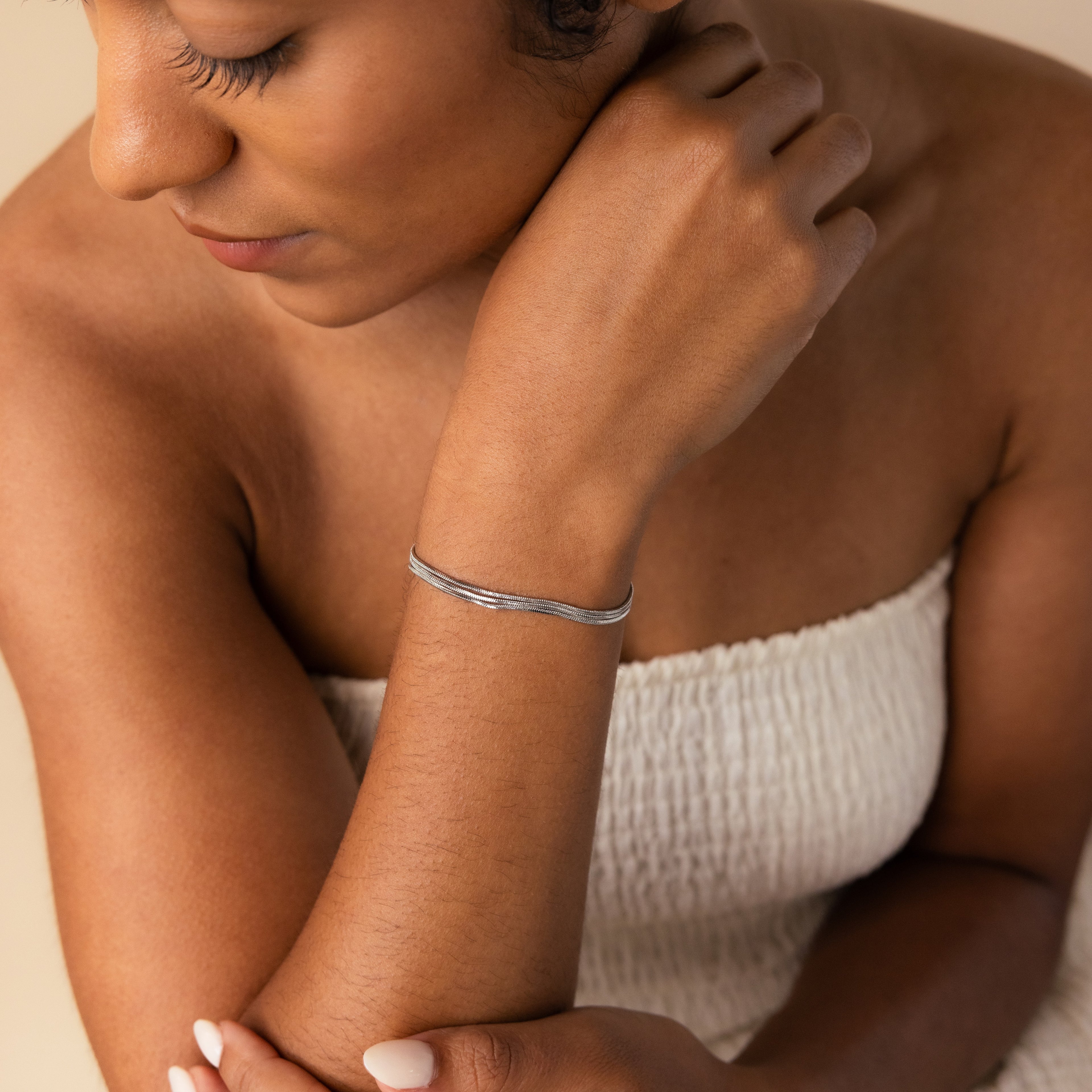 A woman in a white textured top wears minimal jewelry—a sleek Triple Snake Chain Bracelet in White Gold—as she looks down thoughtfully, her hand resting gently on her shoulder.