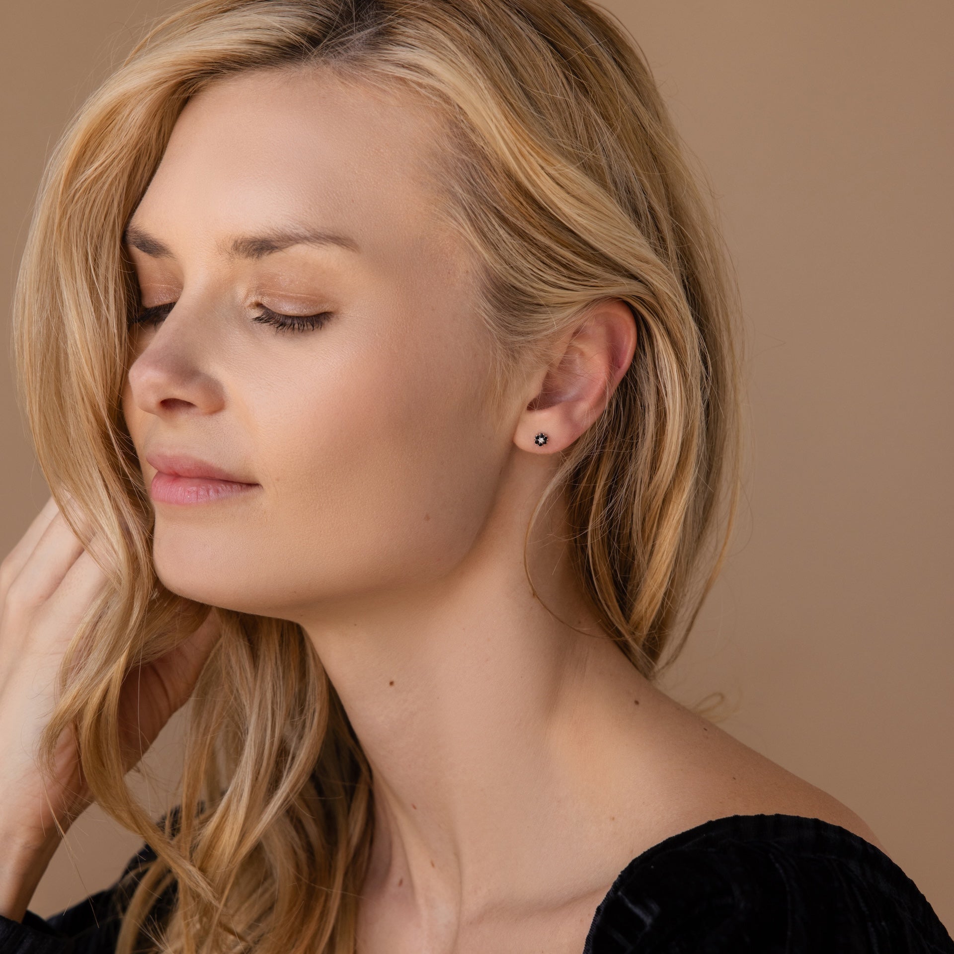 A woman with closed eyes and blonde hair touches her hair, wearing a black top and elegant Onyx Flower Studs in Silver that add a subtle touch of sophistication.