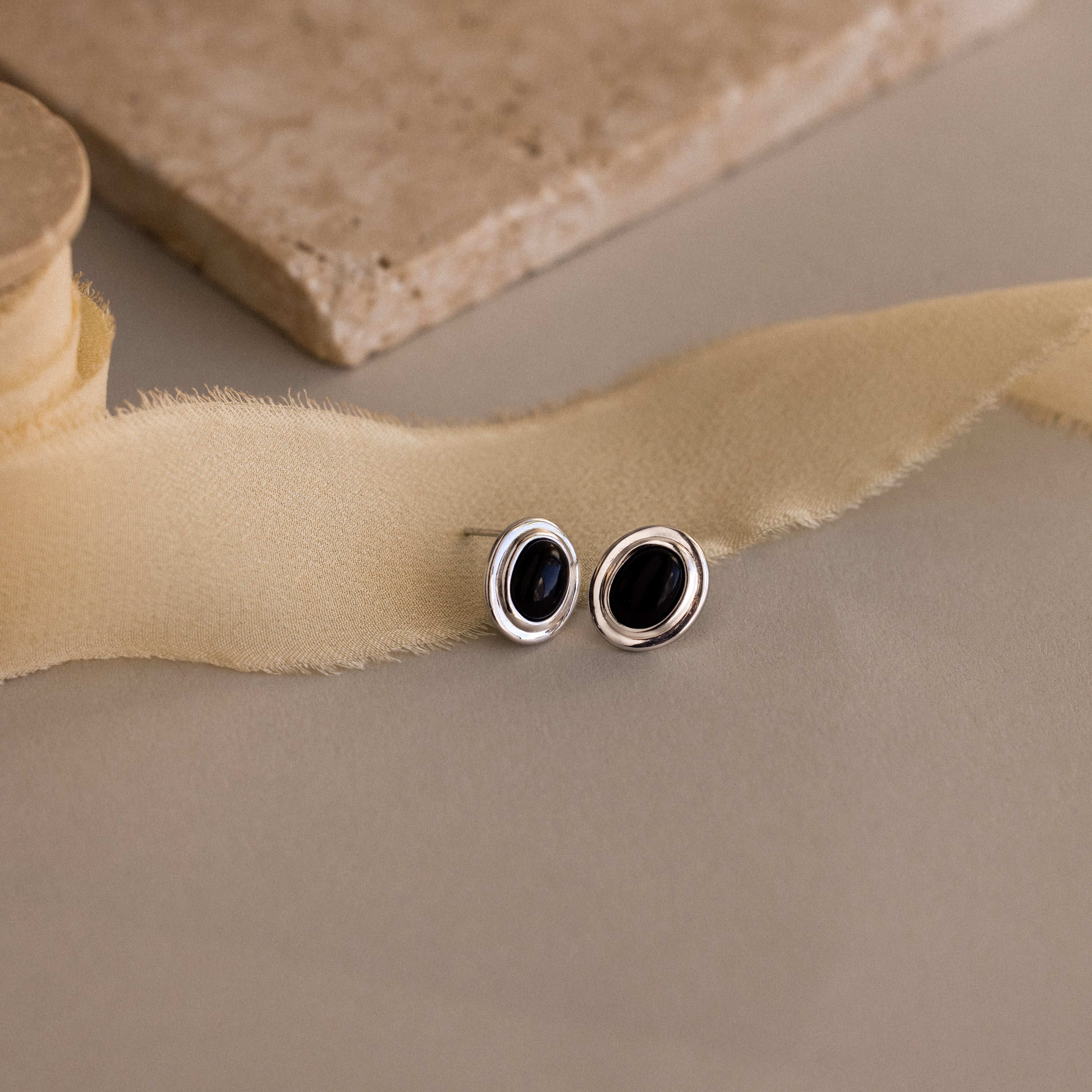 The Vintage Oval Black Studs feature elegant black ovals in silver settings, displayed on a beige surface with fabric and stone accents—perfect for adding a timeless statement to any look.