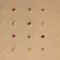 Twelve dainty earrings, including Garnet Flatback Studs in various colors, are arranged in a grid on a beige background.
