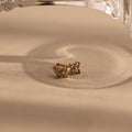A pair of gold-toned stud earrings with four small light purple gemstones arranged in a square, resting on a beige surface with a soft shadow and a glass object partially visible at the top.